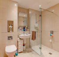 Modern bathroom with beige tiles, a glass-enclosed shower, white sink and toilet, wall shelf with toiletries, and a towel hanging beside the shower.