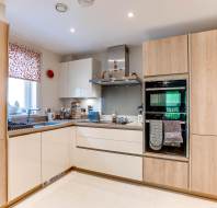 Modern kitchen with light wood and white cabinets, built-in oven, induction cooktop, and various items on the countertops. Window with floral curtain on the left.