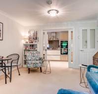 A bright living room with patterned armchairs, a blue sofa, a small round dining table, and a view into a modern kitchen through glass double doors.