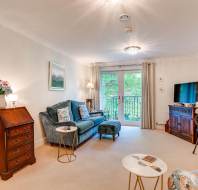 Bright living room with a sofa, TV, small dining table, and French doors leading to a balcony with garden views; traditional and modern furnishings are present.