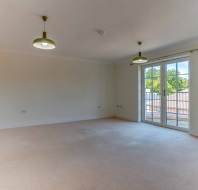 Unfurnished living room with beige carpet, two ceiling lights, large window doors opening to a balcony, and an open doorway leading to the kitchen.