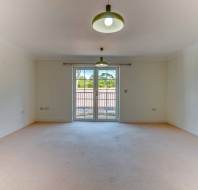 Empty, carpeted living room with two ceiling lights, white walls, and glass doors leading to a balcony with a view of trees.