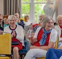 A group of elderly people sit together in a brightly lit room, smiling and chatting; some hold drinks, and one uses a tablet.