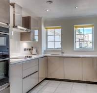Modern kitchen with beige cabinets, built-in ovens, electric cooktop, stainless steel hood, white countertops, sink, and two windows with yellow blinds.