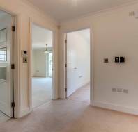 A hallway in a modern home with beige carpet, white walls, two open doors leading to bright rooms, and tile flooring visible in an adjacent area.
