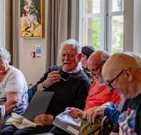 A group of older adults sit together indoors, engaged in conversation and looking at papers, with large windows in the background.