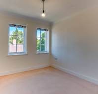 An empty room with beige carpet, cream walls, two windows with patterned valances, and a single exposed ceiling light bulb.