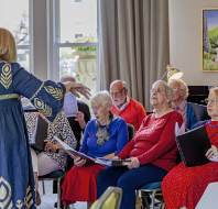 A choir of elderly people sits in a room, holding sheet music, while a woman in a blue dress stands in front, conducting them.