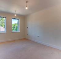 Empty room with beige carpet, cream walls, two ceiling light bulbs, two windows with floral valances, and a view of trees outside.