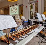A set of handbells is arranged on a table with sheet music. Several people are seated facing a screen displaying a "VE Day" logo in a brightly lit room.