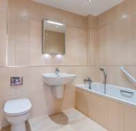 Modern bathroom with beige tiles, a wall-mounted toilet, a sink with a mirror above it, and a bathtub with a grab bar and handheld showerhead.