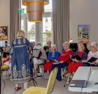 A group of elderly people sit in a semicircle, holding songbooks, as a woman stands in front of them conducting in a well-lit room.