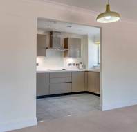 Modern kitchen with beige cabinets, built-in appliances, and tiled floor, viewed through a square opening from a carpeted living area with a single pendant light.