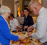 Several people serve themselves scones and cake from a buffet table at an indoor social event. Plates of baked goods are arranged in the foreground.
