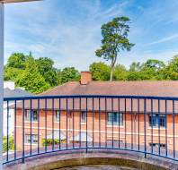 View from an open window showing a curved balcony railing, red-brick building with a pitched roof, and trees in the background under a blue sky.