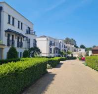A pathway lined with hedges leads past modern white apartment buildings under a clear blue sky.