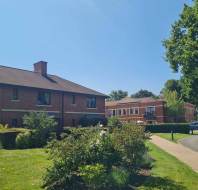 A sunny outdoor scene with red-brick buildings, green bushes, trees, a curved road, and a clear blue sky.