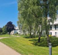A paved path curves through a grassy area with trees, beside a white multi-story building on a clear, sunny day.