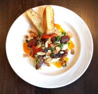 A plated dish featuring sausage pieces, toasted bread slices, sliced almonds, microgreens, sauce, and cheese on a white plate, set on a dark wooden table.