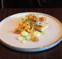 A plated gourmet dish with diced vegetables, a toast topped with a creamy spread, radish slices, green sauce dollops, and microgreens on a white plate.