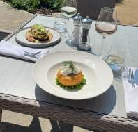 Two plates of gourmet food, two glasses of wine, water, and cutlery are set on an outdoor table in sunlight, with a salt and pepper shaker in the center.
