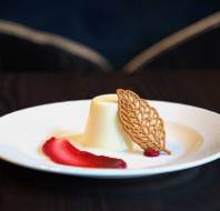 A white dessert, possibly panna cotta, served on a white plate with a decorative cookie and a red fruit sauce smear, set on a dark table.