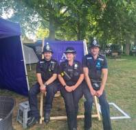 Three police officers in uniform sit on a large wooden chair outdoors under a tree, with tents and park benches visible in the background.