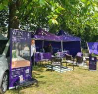 Outdoor promotional booth for Audley Villages with purple tents, banners, seating area, and a parked car under leafy trees on a sunny day.