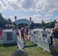 People gather near a river under a decorated archway reading “Jim Campbell Food & Photography”; white fences, lavender plants, and information boards are visible on a sunny day.