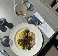 A plated meal with grilled meat, potato, and greens topped with salsa sits on a gray table next to a glass of rosé, water, and a fork with a napkin.