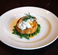 A fish cake topped with a poached egg and creamy sauce, garnished with dill, served on a bed of green peas on a white plate.