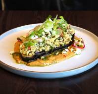 A plated dish featuring roasted eggplant topped with a grain salad, garnished with sliced radishes, greens, and crumbled cheese, served on a smear of sauce.