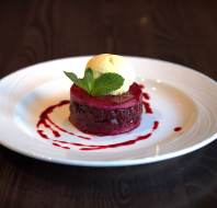 A dessert topped with a scoop of vanilla ice cream and a mint leaf sits on a white plate, drizzled with red sauce, on a dark wooden table.