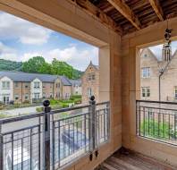 View from a covered balcony with wooden ceiling, overlooking a street with stone houses and greenery in the background under a partly cloudy sky.