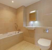 Modern bathroom with beige tiled walls and floor, featuring a bathtub with shower, wall-mounted sink and mirror, and a wall-mounted toilet with a toilet paper holder.