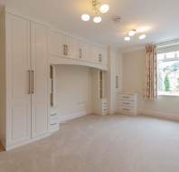 A spacious, carpeted bedroom with built-in white wardrobes, drawers, and a window with beige curtains letting in natural light.