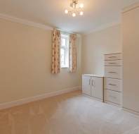 Empty beige carpeted room with a window, floral curtains, and a white wardrobe and drawers unit on the right side. Ceiling light fixture is turned on.