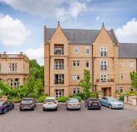 Parking lot with six cars in front of a modern stone apartment building, with an older stone building to the left and greenery around.