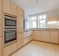 Modern kitchen with light wood and beige cabinets, built-in double oven, electric stove, large sink, tiled floor, and two windows providing natural light.