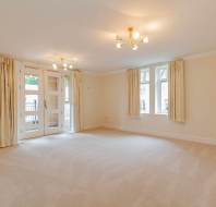 A spacious, empty living room with beige carpet, cream walls, ceiling lights, large windows, and french doors with curtains leading to a balcony.