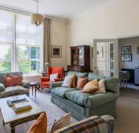 A bright, neatly arranged living room with a sofa, armchairs, coffee table, large windows, and an open doorway leading to a kitchen area.