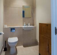 A modern bathroom with a wall-mounted toilet, a small sink, a mirror, soap dispensers, a toilet paper roll, a toilet brush, and a wooden cabinet.