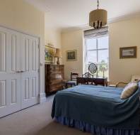 A neatly arranged bedroom with a double bed, wooden furniture, a window with a view, and paintings on light-colored walls.