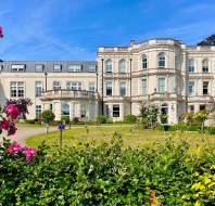 A large white historic building with ornate architecture, surrounded by a manicured garden and blooming flowers under a clear blue sky.