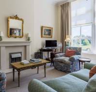 A bright living room with patterned armchairs, a green sofa, a coffee table, a fireplace with a mirror, and large windows overlooking a green landscape.