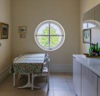 A small kitchen with a floral tablecloth on a dining table, white chairs, a round window, wall art, cabinets, a microwave, and bottles on the counter.