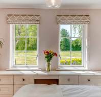 A bedroom with two windows overlooking a green lawn, a vase of flowers on the desk, and a potted plant on the left side.