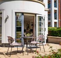 Small patio with two chairs and a round table set outside glass double doors of a modern apartment building, with greenery and other buildings visible.