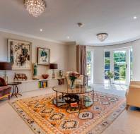 A bright living room with large windows, a patterned rug, two armchairs, a glass coffee table, bookshelves, and wall art.