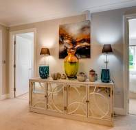 A hallway with a decorative sideboard, vases, lamps, and artwork, leading to a living room on the left and a bedroom on the right with large windows.